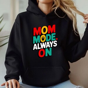 May include: A black hooded sweatshirt featuring the text "MOM MODE. ALWAYS ON" printed on the chest. The word "MOM" is in red, "MODE" is in teal with a yellow power symbol, and "ALWAYS ON" is in white and red with teal accents.