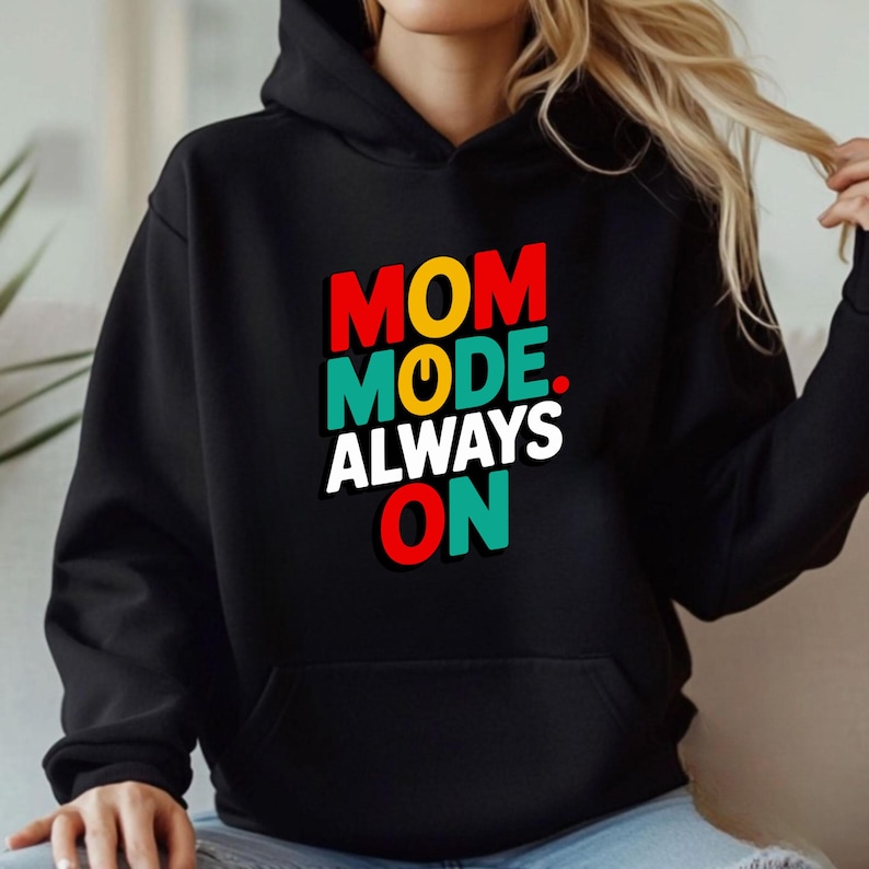 May include: A black hooded sweatshirt featuring the text "MOM MODE. ALWAYS ON" printed on the chest. The word "MOM" is in red, "MODE" is in teal with a yellow power symbol, and "ALWAYS ON" is in white and red with teal accents.