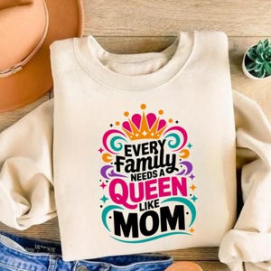 May include: A cream-colored crewneck sweatshirt featuring a colorful graphic print that reads Every Family Needs A Queen Like Mom in black, pink, and orange lettering, accented by a stylized crown and swirling decorative flourishes.