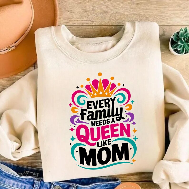 May include: A cream-colored crewneck sweatshirt featuring a colorful graphic print that reads Every Family Needs A Queen Like Mom in black, pink, and orange lettering, accented by a stylized crown and swirling decorative flourishes.