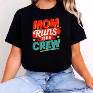 May include: A black short-sleeved t-shirt featuring a graphic print with the text "MOM RUNS THIS CREW" in red, white, and teal lettering. The design includes small yellow star accents and is centered on the chest of the garment.
