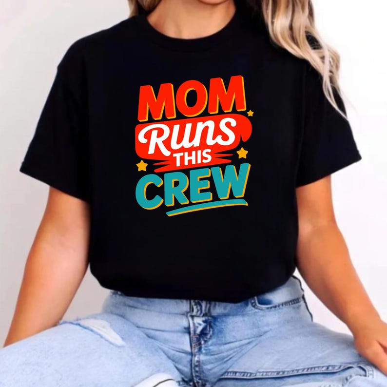 May include: A black short-sleeved t-shirt featuring a graphic print with the text "MOM RUNS THIS CREW" in red, white, and teal lettering. The design includes small yellow star accents and is centered on the chest of the garment.