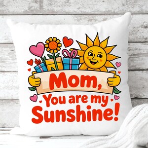 May include: A white square throw pillow featuring a colorful graphic of a smiling yellow sun, a sunflower, and gift boxes. The text reads, "Mom, You are my Sunshine!" surrounded by small pink and red hearts on a light-colored fabric surface.