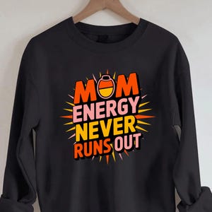 May include: A black long-sleeve sweatshirt featuring a graphic design with the text "MOM ENERGY NEVER RUNS OUT" in orange, pink, and yellow. The letter O in MOM is replaced by a candy corn icon. The design is surrounded by a sunburst pattern.