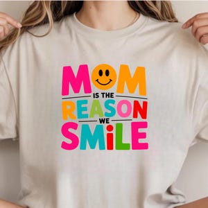 May include: A beige short-sleeved t-shirt featuring a multi-colored graphic print. The text reads "MOM IS THE REASON WE SMILE" in bold, vibrant letters, with a yellow smiley face replacing the letter "O" in the word "MOM" at the top center.