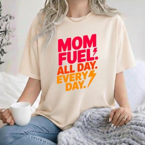 May include: A cream-colored short-sleeve t-shirt featuring the red and orange text "MOM FUEL. ALL DAY. EVERY DAY." with a lightning bolt graphic. The shirt has a crew neckline and a relaxed fit, displaying bold typography on the front chest area.
