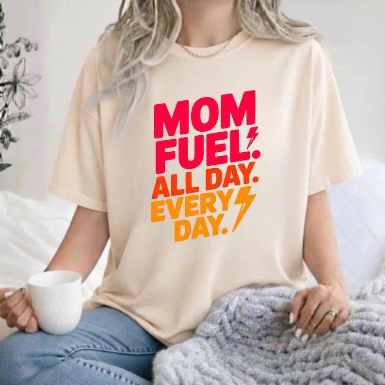 May include: A cream-colored short-sleeve t-shirt featuring the red and orange text "MOM FUEL. ALL DAY. EVERY DAY." with a lightning bolt graphic. The shirt has a crew neckline and a relaxed fit, displaying bold typography on the front chest area.