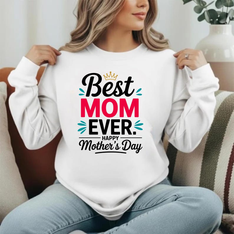 May include: A white long-sleeve sweatshirt featuring a graphic print with the text "Best MOM EVER. HAPPY Mother's Day" in black, red, and teal. The design includes a small gold crown icon above the word "Best" and decorative teal accents.