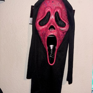 May include: A full-head costume mask featuring a bright pink face with dark purple paint splatters, large black eye openings, and an elongated open mouth. A small gold-colored hoop ring is attached to the nose area, and a black fabric hood is attached.