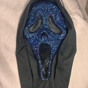 May include: A ghost-style mask featuring a face covered in iridescent blue and purple rhinestones, with a black rhinestone-encrusted mouth area and black mesh eye openings. The mask is attached to a plain black fabric hood that drapes downwards.