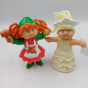 May include: Two small plastic dolls with yarn hair. One doll has orange hair, a green hat, a red and white apron with a bear, and holds a brown mallet. The other doll has blonde hair, a white crown, and a glittery white dress with a snowflake.