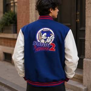 May include: A blue varsity jacket with white sleeves and red and blue striped ribbed trim at the collar, cuffs, and hem. The back features a large embroidered patch of Sonic the Hedgehog with the text Sonic The Hedgehog 2 in red and blue.