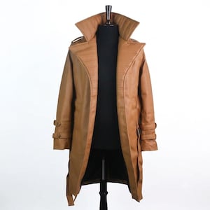 May include: A tan trench coat featuring a wide, structured collar with horizontal stitching, long sleeves with adjustable buckled straps at the cuffs, and a long, open-front design. The coat is displayed on a black mannequin torso.