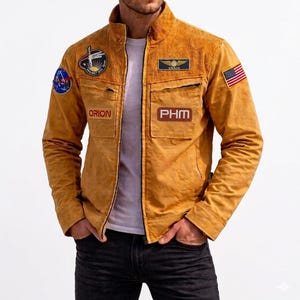 Puede incluir: Una chaqueta de pana color amarillo mostaza con cuello alzado, cremallera frontal y varios parches, incluyendo un logo de la NASA, una bandera de EE. UU. y etiquetas rectangulares con el texto ORION y PHM, sobre una camiseta blanca.