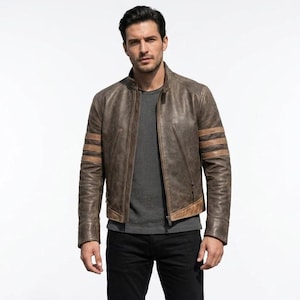 Può includere: Una giacca in pelle marrone effetto invecchiato con colletto alla coreana, chiusura frontale con zip e tre strisce orizzontali beige su ogni manica. La giacca è indossata sopra una t-shirt grigia tinta unita e abbinata a pantaloni neri su sfondo bianco.