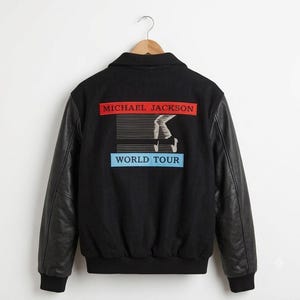 May include: A black varsity jacket with black leather sleeves and a ribbed collar, cuffs, and hem. The back features a graphic with the text MICHAEL JACKSON in a red rectangle, a white illustration of legs, and the text WORLD TOUR in a blue rectangle.