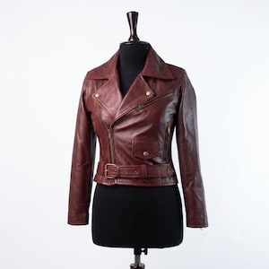 May include: A deep burgundy leather motorcycle jacket featuring an asymmetrical front zip, a wide notched collar, long sleeves, and a buckled waist belt. The jacket includes a small flap pocket on the front and additional zipped pockets.