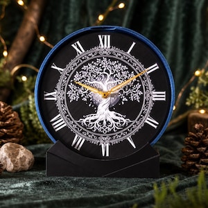 Può includere: Un orologio nero e blu con un design ad albero della vita bianco. Il quadrante presenta numeri romani e lancette dorate. L'orologio è su un supporto nero, con pigne e vegetazione sullo sfondo.