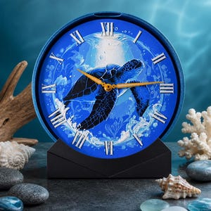 May include: A blue clock with a sea turtle design. The clock face features a detailed illustration of a turtle in shades of blue, surrounded by Roman numerals. The clock has gold-colored hands and sits on a black stand.