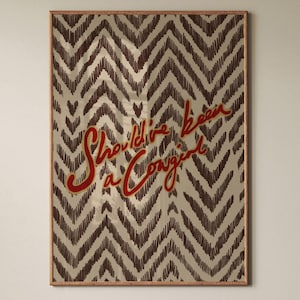 May include: A rectangular wall art print featuring a dark brown chevron pattern on a light beige background. The text "Should've been a cowgirl" is written in a bold, red, cursive script across the center of the chevron design.