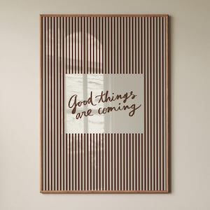 Può includere: Una stampa artistica da parete rettangolare in una cornice di legno con un motivo a strisce verticali marrone scuro. Un'area bianca rettangolare centrale mostra il testo scritto a mano in marrone scuro Good things are coming sullo sfondo a righe.