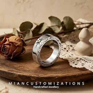 May include: A silver-toned metal ring featuring a polished finish with an engraved sunburst motif centred on a small gold-coloured dot. The band is decorated with black etched swirling patterns and dots along its outer circumference.