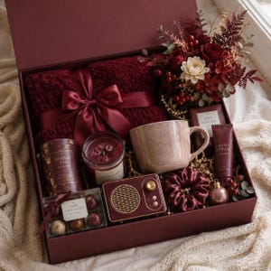 Op de afbeelding: Een geschenkdoos met een donkerrode gebreide deken met satijnen lint, een bloemstuk, een gespikkelde keramische mok, een kaars, kruidenthee, pure chocoladetruffels, een vintage-stijl luidspreker, een scrunchie en handcrème.