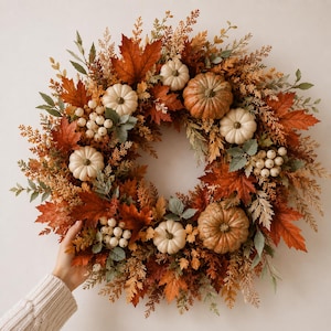 Peut inclure: Une couronne d'automne circulaire composée d'un mélange de feuilles d'érable orange, rouges et brunes, entremêlées de petites citrouilles blanches, de plus grosses citrouilles orange texturées et de grappes de petites baies blanches.