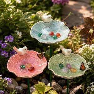 Puede incluir: Tres bebederos para pájaros de cerámica en un jardín. Cada bebedero tiene forma de hoja y presenta una pequeña figura de pájaro blanco en la parte superior. Los bebederos son de color rosa pastel, verde y azul, y contienen piedras de vidrio de colores.