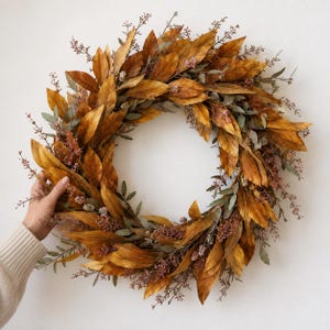 Puede incluir: Una corona circular compuesta por hojas otoñales de color marrón dorado, intercaladas con follaje verde apagado y delicados racimos de pequeñas bayas de color rosa parduzco. El arreglo presenta una textura densa y estratificada.