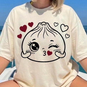 May include: A cream-colored t-shirt featuring a black line-art graphic of a winking dumpling character. The dumpling has a kissy face, blushing cheeks, and is surrounded by several small red and black heart icons printed on the front center.