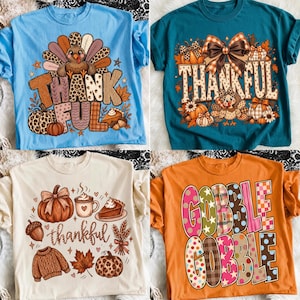 May include: Four t-shirts featuring Thanksgiving-themed graphics. The top left is light blue with a turkey and the word THANKFUL. The top right is teal with a turkey, bow, and THANKFUL. The bottom left is cream with autumn icons and thankful. The bottom right is orange with GOBBLE GOBBLE.
