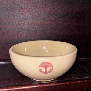 May include: A small, round, cream-colored ceramic bowl with a smooth, glossy finish. Centered on the front exterior is a circular red emblem featuring a caduceus symbol and the text "UNITED STATES PUBLIC HEALTH SERVICE".