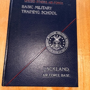 May include: A navy blue book with embossed text reading "UNITED STATES AIR FORCE BASIC MILITARY TRAINING SCHOOL" and "LACKLAND AIR FORCE BASE." A red diagonal line accents the cover, and a circular emblem is present.