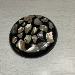 May include: A round, black decorative object inlaid with iridescent, irregularly shaped pieces. The pieces have a pearlescent sheen, reflecting colors like silver, gold, and green. The object is set against a light-colored wooden surface.