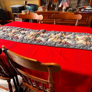 May include: A red tablecloth with a quilted table runner. The runner is rectangular with a gray border and a patchwork design in shades of beige, red, and green. Wooden chairs surround the table.