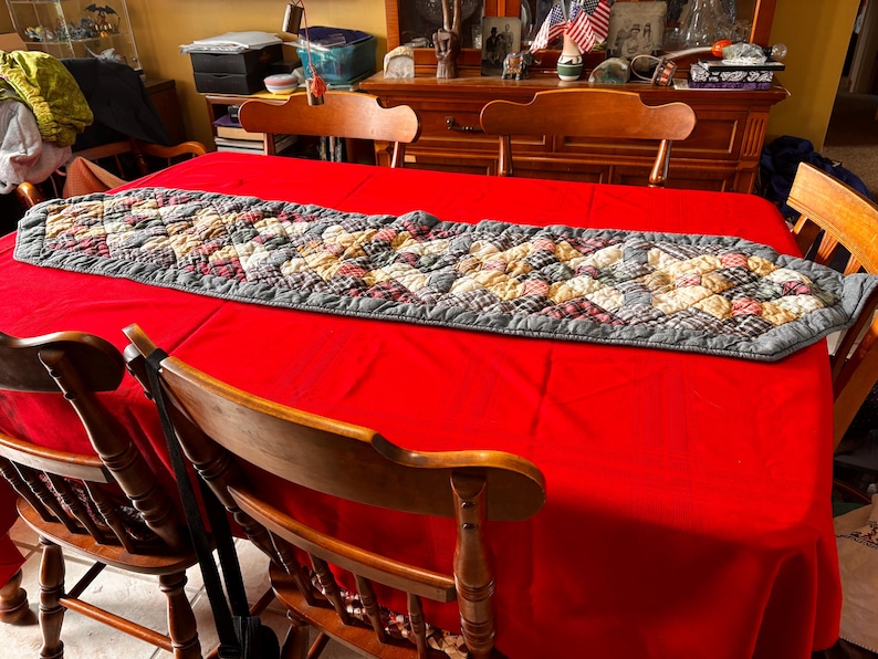 K&ouml;nnte beinhalten: Eine rote Tischdecke mit einem gesteppten Tischl&auml;ufer. Der L&auml;ufer hat einen grauen Rand und ein Patchwork-Design in Beige-, Rot- und Gr&uuml;nt&ouml;nen. Holzst&uuml;hle umgeben den Tisch.