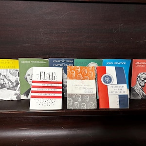 Może przedstawiać: Kolekcja siedmiu małych, kolorowych książek o historii Ameryki. Książki przedstawiają Deklarację Niepodległości, George'a Washingtona, Konstytucję Stanów Zjednoczonych, patriotyczne pieśni Ameryki, Johna Hancocka i Abrahama Lincolna. Książki są ułożone w rzędzie na ciemnym drewnianym podłożu.