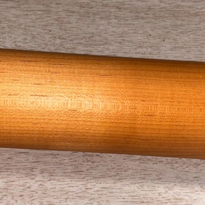 May include: A light brown wooden rolling pin with rounded handles. The rolling pin is made of a light-colored wood and has a smooth, polished finish. The handles are teardrop-shaped. A small logo is visible on the rolling pin.