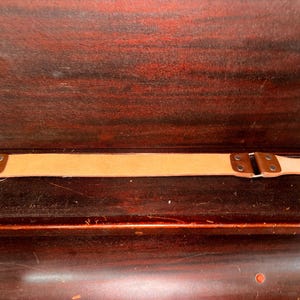 Puede incluir: Un cuero de afilado de cuero color canela con detalles de cuero marrón y un mango de madera. El cuero, utilizado para afilar navajas rectas, tiene un cierre metálico para colgar. Mide unos 50 cm.