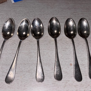 Puede incluir: Seis cucharas de plata con cuencos ovalados y mangos largos y delgados están dispuestas sobre una superficie de color claro. Las cucharas parecen ser vintage o antiguas, con un acabado ligeramente deslustrado. Los mangos tienen un diseño grabado sutil.