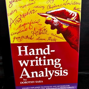 Op de afbeelding: Een boek getiteld "Handwriting Analysis" door Dorothy Sara. De omslag is rood met witte tekst en een gele achtergrond met woorden als "Agressief" en "Artistiek". Een hand die een pen vasthoudt, is afgebeeld.
