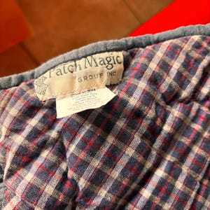 K&ouml;nnte beinhalten: Nahaufnahme eines gesteppten Stoffes mit einem Karomuster in Marineblau, Rot und Wei&szlig;. Der Stoff hat ein Etikett mit der Aufschrift "Patch Magic GROUP INC". Der Stoff ist wahrscheinlich Teil einer Steppdecke oder eines &auml;hnlichen Textils.