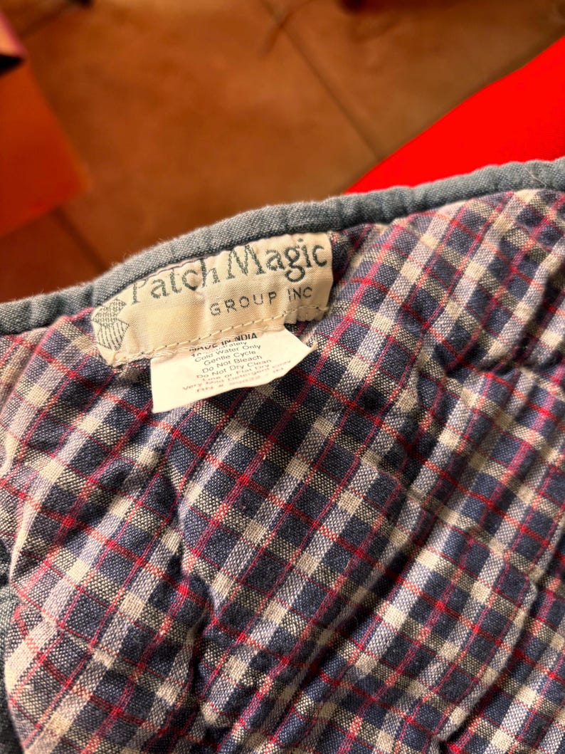 K&ouml;nnte beinhalten: Nahaufnahme eines gesteppten Stoffes mit einem Karomuster in Marineblau, Rot und Wei&szlig;. Der Stoff hat ein Etikett mit der Aufschrift "Patch Magic GROUP INC". Der Stoff ist wahrscheinlich Teil einer Steppdecke oder eines &auml;hnlichen Textils.
