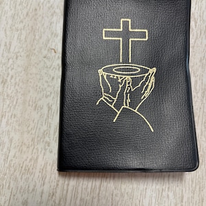 May include: Black leather-bound book with a gold cross and chalice design. The cover features a textured surface and a simple, elegant design. The book appears to be a religious text, possibly a Bible or prayer book.