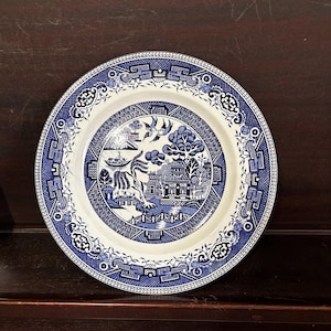 May include: A blue and white ceramic plate with a traditional willow pattern design. The plate features a stylized willow tree, a pagoda, and a bird in flight.