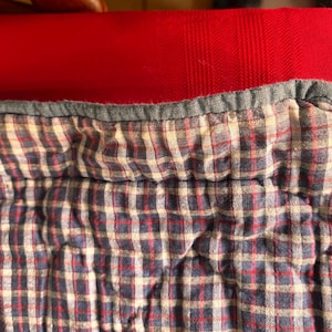 K&ouml;nnte beinhalten: Nahaufnahme eines gesteppten Stoffes mit einem blau-rot-wei&szlig; karierten Muster. Der Stoff hat einen grauen Rand und ist vor einem roten Hintergrund platziert. Die Textur des Stoffes ist weich und leicht zerknittert.