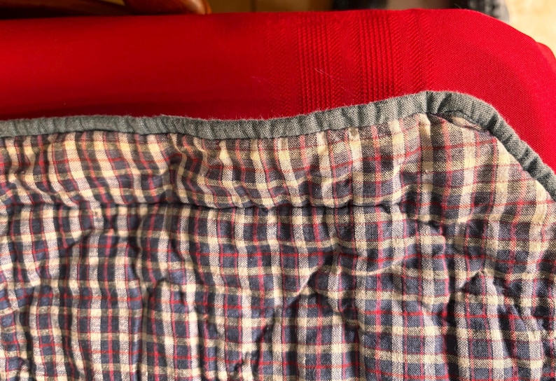 K&ouml;nnte beinhalten: Nahaufnahme eines gesteppten Stoffes mit einem blau-rot-wei&szlig; karierten Muster. Der Stoff hat einen grauen Rand und ist vor einem roten Hintergrund platziert. Die Textur des Stoffes ist weich und leicht zerknittert.