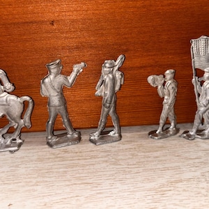 May include: A set of small, silver toy soldiers. The figures represent soldiers in different poses, including a rider on horseback, a soldier aiming a weapon, and soldiers carrying flags and musical instruments. The figures are set against a wooden background.