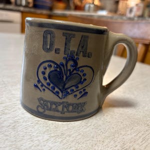 May include: A brown ceramic mug with blue glaze. It has a heart design with blue dots and the text "O.T.A. Salt Fork" on the front.